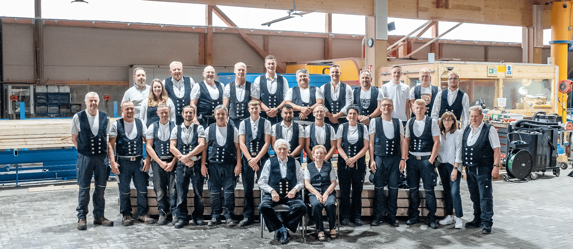 Gruppe von 30 Personen in traditioneller Kleidung, die in einer Werkstatt stehen und sitzen. Einige Personen tragen schwarze Westen mit weißen Hemden.